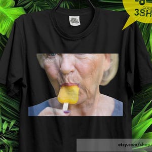 Könnte beinhalten: Ein schwarzes Kurzarm-T-Shirt mit einem mittigen Fotodruck einer Person, die ein gelbes Eis am Stiel isst. Das Kleidungsstück hat einen Rundhalsausschnitt und ist auf einem schwarzen Kunststoffbügel vor grünen Tropenblättern abgebildet.