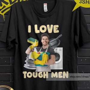 May include: A black short-sleeved t-shirt featuring a graphic print of a person wearing yellow rubber gloves and holding a blue bucket filled with cleaning supplies. The text I LOVE TOUGH MEN is printed in yellow above and below the central image.