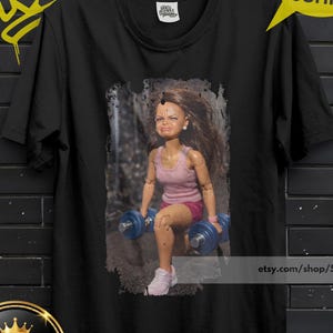 Könnte beinhalten: Schwarzes T-Shirt mit einer Puppe, die Gewichte hebt, ein rosa Tanktop und Shorts trägt. Das Shirt hat den Text "Sub Urban" im gelben Graffiti-Stil. Eine gelbe Sprechblase sagt "KAUFEN 3 KOSTENLOSER VERSAND" mit dem Code "3SHIP".