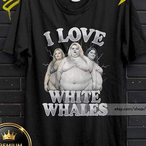 May include: A black short-sleeve t-shirt featuring a graphic print of three individuals with the text I LOVE WHITE WHALES in a bold, silver-toned font. The design includes a lightning bolt background effect behind the figures on the front of the shirt.