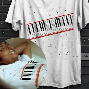 Puede incluir: Una camiseta blanca de manga corta con un gráfico de teclado de piano en blanco y negro colocado en diagonal sobre el pecho, acentuado por dos finas líneas horizontales rojas y notas musicales negras dispersas sobre y bajo las teclas.