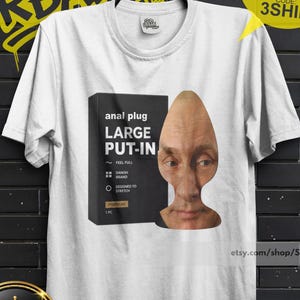 Może przedstawiać: Biały t-shirt z krótkim rękawem z nadrukiem graficznym przedstawiającym czarne pudełko z napisem 'anal plug LARGE PUT-IN' obok ludzkiej twarzy w kształcie korka. Koszulka ma okrągły dekolt i jest zawieszona na czarnym wieszaku.