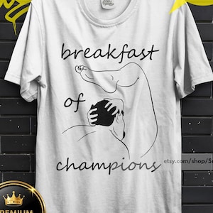 Könnte beinhalten: Ein weißes Kurzarm-T-Shirt mit einer schwarzen Strichzeichnung, die eine Person bei der Oralverkehr-Ausübung zeigt. Der Text "breakfast of champions" ist in einer schwarzen Schreibschrift über und unter der Illustration aufgedruckt.