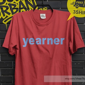 Op de afbeelding: Rood t-shirt met het woord "yearner" in lichtblauwe letters. Het t-shirt hangt aan een zwarte hanger tegen een zwarte bakstenen muur. Een gele sticker met "BUY 3 FREE SHIP" staat rechtsboven.