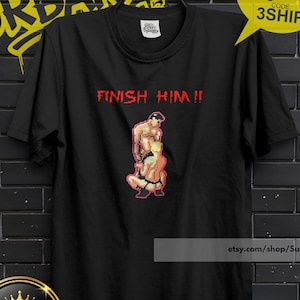 Könnte beinhalten: Schwarzes T-Shirt mit dem roten Text "FINISH HIM!!". Das Shirt zeigt eine pixelige Grafik von zwei Figuren in Kampfpose. Eine gelbe Sprechblase mit "BUY 3 FREE" und "CODE: 3SHIP" befindet sich rechts.