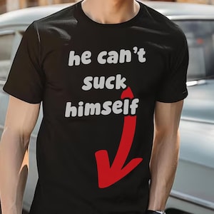 Op de afbeelding: Een zwart t-shirt met korte mouwen en de witte tekst "he can't suck himself" op de borst, vergezeld van een grote, opvallende rode pijl die naar beneden wijst richting de onderkant van het kledingstuk.