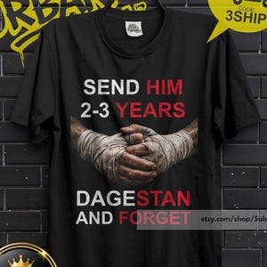 May include: A black short-sleeved t-shirt featuring a graphic of hands wrapped in white boxing tape. The text reads SEND HIM 2-3 YEARS in white and red, and DAGESTAN AND FORGET in white and red. The shirt is displayed on a black hanger.