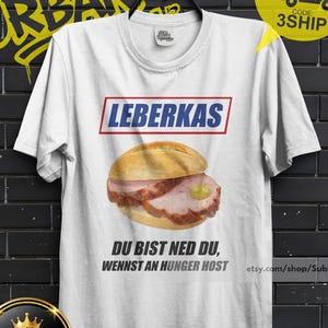 May include: White t-shirt featuring a graphic of a Leberkas sandwich with the word "LEBERKAS" above it. The shirt also has text that reads "DU BIST NED DU, WENNST AN HUNGER HOST". A yellow circle says "BUY 3 FREE" and "3SHIP".