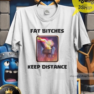 Può includere: T-shirt bianca con la scritta "FAT BITCHES KEEP DISTANCE" in nero. La maglietta presenta una grafica di un personaggio di un videogioco. La maglietta è esposta su una gruccia su uno sfondo blu e oro.