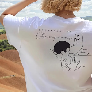 May include: White t-shirt with the text "BREAKFAST OF CHAMPIONS" in a script font. The back of the shirt features a black and white line drawing of a person's head and arm.