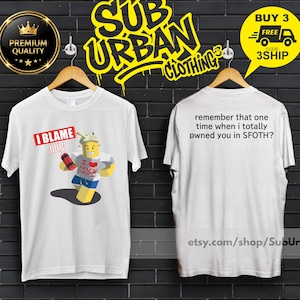 Può includere: T-shirt bianche con grafiche. Una maglietta presenta un personaggio dei cartoni animati con la scritta "I BLAME SOMEONE" e "I FRIED CHICKEN". L'altra maglietta ha la scritta "remember that one time when I totally pwned you in SFOTH?"