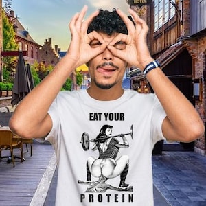 Könnte beinhalten: Ein weißes T-Shirt mit dem Text "EAT YOUR PROTEIN" und einer Grafik einer Frau, die Gewichte hebt. Die Person auf dem Bild macht eine "okay"-Geste mit ihren Händen über ihren Augen.