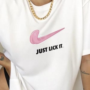Può includere: Una t-shirt bianca a maniche corte con la grafica di una lingua rosa a forma di logo swoosh. Sotto la grafica, il testo "JUST LICK IT." è stampato in lettere maiuscole nere in grassetto. La maglietta ha un girocollo e una vestibilità ampia.
