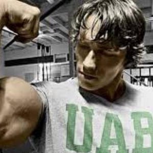 Könnte beinhalten: Eine Person, die ihren Bizeps anspannt. Die Person trägt ein graues T-Shirt mit den grünen Buchstaben "UAB" auf der Vorderseite. Der Hintergrund ist ein Fitnessstudio mit Trainingsgeräten.