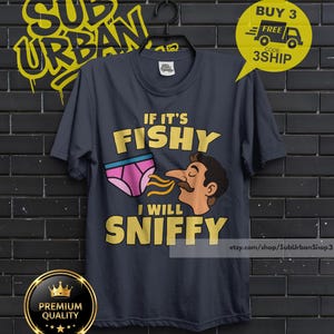 May include: A dark gray short-sleeve t-shirt featuring a graphic of a person with a mustache sniffing a pair of pink underwear. The text on the shirt reads IF IT'S FISHY I WILL SNIFFY in bold yellow letters. The shirt is displayed on a black hanger.
