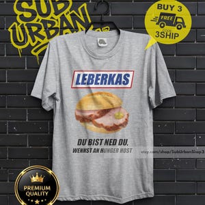 May include: A gray t-shirt featuring a Leberkas sandwich graphic with the text "LEBERKAS" above and "DU BIST NED DU, WENNST AN HUNGER HOST" below. The shirt also has a "Premium Quality" badge.