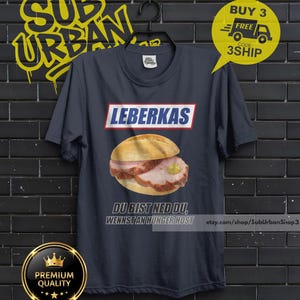 May include: Navy blue t-shirt featuring a Leberkas sandwich illustration. The word "LEBERKAS" is printed above the sandwich, with the phrase "DU BIST NED DU, WENNST AN HUNGER HOST" below. The shirt also has a "BUY 3 FREE" promotion.