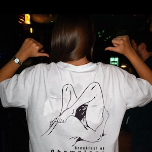 Könnte beinhalten: Weißes T-Shirt mit einer schwarzen Strichzeichnung einer liegenden Figur. Der Text "Breakfast of Champions" ist in Schwarz unter der Grafik aufgedruckt. Das T-Shirt wird von einer Person getragen.