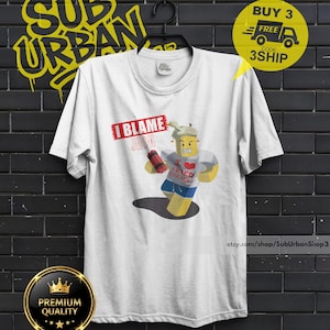 Può includere: T-shirt bianca con un personaggio dei cartoni animati che tiene in mano un oggetto rosso e indossa una maglietta con la scritta "I Love Fried Chicken". Il testo "I Blame Jim" è sopra il personaggio. La maglietta ha un girocollo e maniche corte.