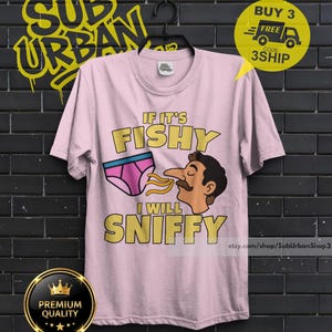 May include: A light pink short-sleeve t-shirt featuring a graphic of a person with a mustache sniffing a pair of pink underwear. The text reads IF IT'S FISHY I WILL SNIFFY in yellow block letters. The shirt has a crew neckline and a standard fit.