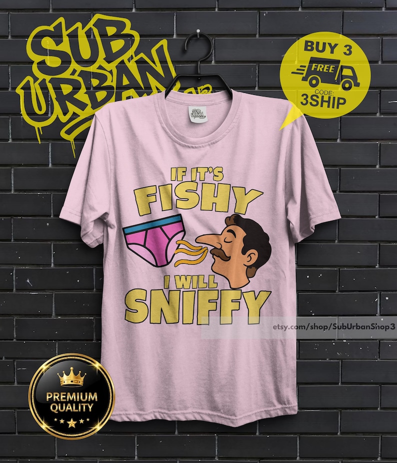 May include: A light pink short-sleeve t-shirt featuring a graphic of a person with a mustache sniffing a pair of pink underwear. The text reads IF IT'S FISHY I WILL SNIFFY in yellow block letters. The shirt has a crew neckline and a standard fit.