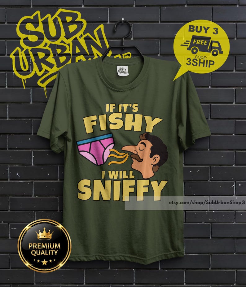 May include: An olive green t-shirt featuring a graphic of a person sniffing a pair of pink underwear. The text reads IF IT'S FISHY I WILL SNIFFY in bold yellow letters. The shirt has a crew neck and short sleeves, displayed on a black hanger.