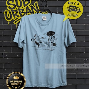 May include: A light blue short-sleeve t-shirt featuring a black graphic print of a cartoon character running past a tree and another character. The shirt has a crew neckline and a small white brand label on the inside of the collar.