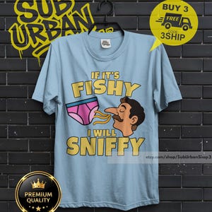 May include: A light blue t-shirt featuring a graphic of a person with a mustache sniffing a pair of pink underwear. The text reads IF IT'S FISHY I WILL SNIFFY in yellow letters. The shirt is displayed on a black hanger against a dark brick wall.