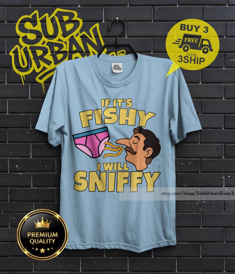May include: A light blue t-shirt featuring a graphic of a person with a mustache sniffing a pair of pink underwear. The text reads IF IT'S FISHY I WILL SNIFFY in yellow letters. The shirt is displayed on a black hanger against a dark brick wall.