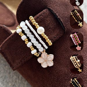 Könnte beinhalten: Eine Kollektion von mehrreihigen Perlenarmbändern mit verschiedenen Anhängern, darunter eine rosa Blume, eine Muschel, eine Schildkröte und ein Seestern. Die Armbänder bestehen aus kleinen metallischen, weißen, rosa, blauen und schwarzen Perlen.