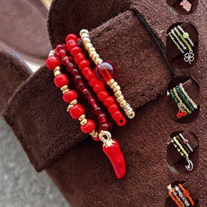 Könnte beinhalten: Ein Set aus drei gestapelten Perlenringen mit einem roten Chilischoten-Anhänger. Die Ringe bestehen aus kleinen roten und goldfarbenen Perlen. Fünf weitere Ring-Sets sind in kreisförmigen Einsätzen dargestellt, mit Anhängern wie Herz, Kleeblatt, Kirschen, Mond und Schmetterling.