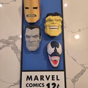 Op de afbeelding: Een ingelijste display met vier superheldenmaskers: Iron Man, Hulk, een grijskleurig personage en Venom. De maskers zijn gemonteerd op een blauwe achtergrond, met een wit bordje onderaan met de tekst "MARVEL COMICS GROUP 12¢".