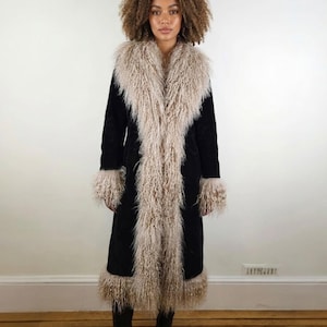 May include: A long black coat with a cream-colored faux fur collar, cuffs, and trim. The coat is knee-length and has a classic silhouette. The model is wearing black boots. This coat is perfect for a vintage or bohemian style.