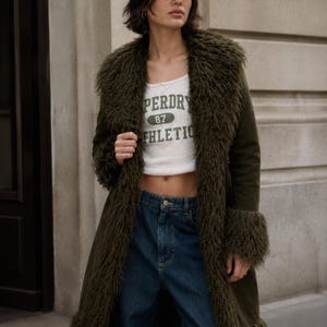 Puede incluir: Un abrigo verde oliva con cuello y puños de piel sintética. La modelo lleva un top corto blanco con el texto "PERDRY 87 ATHLETIC" y una gorra de béisbol verde. La modelo también lleva vaqueros azules.