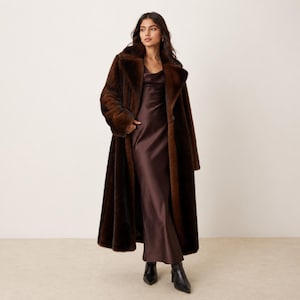 May include: A long, dark brown faux fur coat with wide lapels worn over a matching dark brown satin slip dress that reaches the ankles. The ensemble is completed with a pair of black pointed-toe ankle boots with a block heel, creating a layered look.