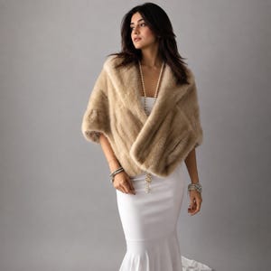 Op de afbeelding: Een beige imitatiebont stola gedrapeerd over een witte mouwloze jurk. De stola heeft een dikke, pluche textuur en brede revers. Een lange streng witte parels hangt om de hals en een breed, meerlaags parelarmband wordt om de pols gedragen.