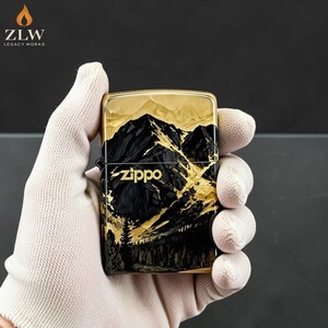 Może przedstawiać: Zapalniczka Zippo z motywem g&oacute;rskiego krajobrazu. Zapalniczka ma czarno-złotą kolorystykę, ze szczeg&oacute;łowym przedstawieniem g&oacute;r i drzew. Logo Zippo jest widoczne z przodu. Zapalniczka jest trzymana przez dłoń w rękawicy.