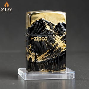 Może przedstawiać: Zapalniczka Zippo z motywem g&oacute;rskiego krajobrazu. Zapalniczka ma czarno-złotą kolorystykę, ze szczeg&oacute;łowym przedstawieniem g&oacute;r i drzew. Logo Zippo jest wyraźnie widoczne. Zapalniczka stoi na przezroczystym stojaku wystawowym.
