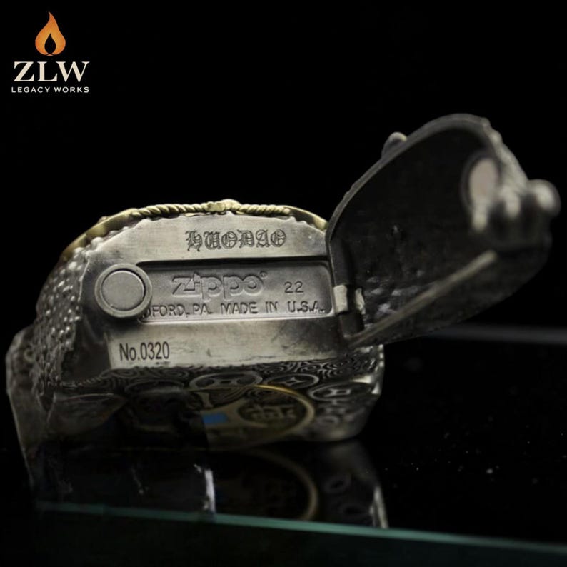 Może przedstawiać: Srebrna zapalniczka Zippo ze skomplikowanymi wytłoczonymi detalami i otwartą pokrywą. Zapalniczka posiada logo Zippo, napis "FORD, PA. MADE IN U.S.A." oraz numer "22". Widoczny jest r&oacute;wnież numer seryjny "No.0320".