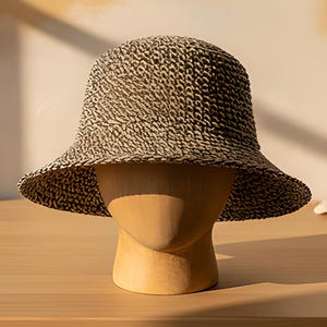 May include: A bucket hat featuring a textured, woven pattern in alternating shades of black and beige. The hat has a rounded crown and a downward-sloping brim, displayed on a smooth, light-toned wooden mannequin head form.