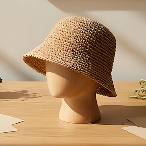May include: A light brown, woven bucket hat displayed on a wooden mannequin head. The hat has a wide brim and a textured appearance. The mannequin is on a wooden table, with a magnifying glass and sketches nearby. The overall theme is fashion and accessories.