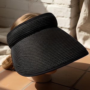 Può includere: Un cappello con visiera nero con una tesa larga, realizzato in materiale tessuto. Il cappello è progettato per proteggere il viso dal sole. La visiera è mostrata su una forma di testa, con un muro di mattoni e tessuto sullo sfondo.
