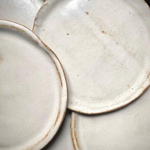 Pode incluir: Um conjunto de pratos de cerâmica esbranquiçada com uma textura salpicada e uma borda marrom rústica. Os pratos têm um design simples e minimalista, perfeitos para servir alimentos. Os pratos são feitos à mão.