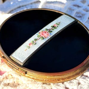 May include: An oval compact case with a polished black surface and a central silver-toned bar featuring a delicate hand-painted floral motif of pink roses and green leaves. The case has a textured gold-toned metal rim with intricate detailing.