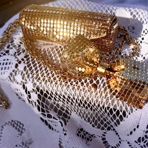 Pode incluir: Uma bolsa clutch de malha metálica dourada com corpo retangular e um laço decorativo na parte frontal. A bolsa inclui uma alça de corrente dourada a condizer, sendo toda a superfície construída com pequenas placas metálicas refletoras.