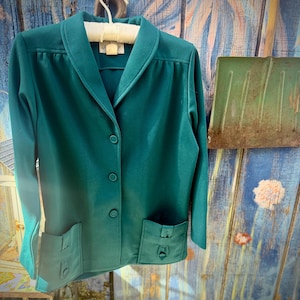 May include: A teal green blazer with a notched collar, three buttons, and two front pockets. The blazer is hanging on a white hanger against a weathered blue and green wooden background. The blazer has a small label on the collar.