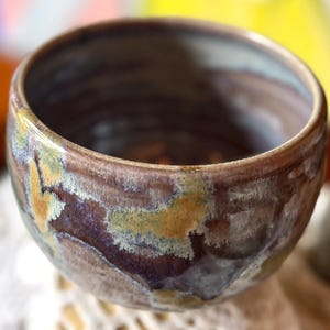 Puede incluir: Un cuenco de cerámica pequeño y redondo con un acabado esmaltado grueso. El exterior presenta un patrón moteado de tonos marrón oscuro, beige terroso y gris apagado. El borde es liso y redondeado, dando paso a un interior profundo.