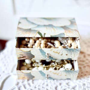 May include: A two-drawer jewelry box featuring a floral pattern in shades of light blue, white, and gold. Each drawer has a small silver knob and is filled with various necklaces, bracelets, and beads in white, cream, and metallic gold tones.