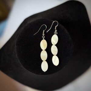 May include: A pair of dangle earrings with three oval, off-white shell pieces suspended from silver-colored hooks. The earrings are displayed on a black, velvet-like surface, showcasing their delicate design and natural color.