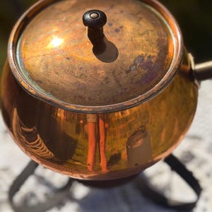 May include: A polished copper pot with a rounded body and a matching lid featuring a small black knob handle. The pot is set upon a dark metal stand with curved legs. The reflective surface of the copper shows distorted outdoor light reflections.